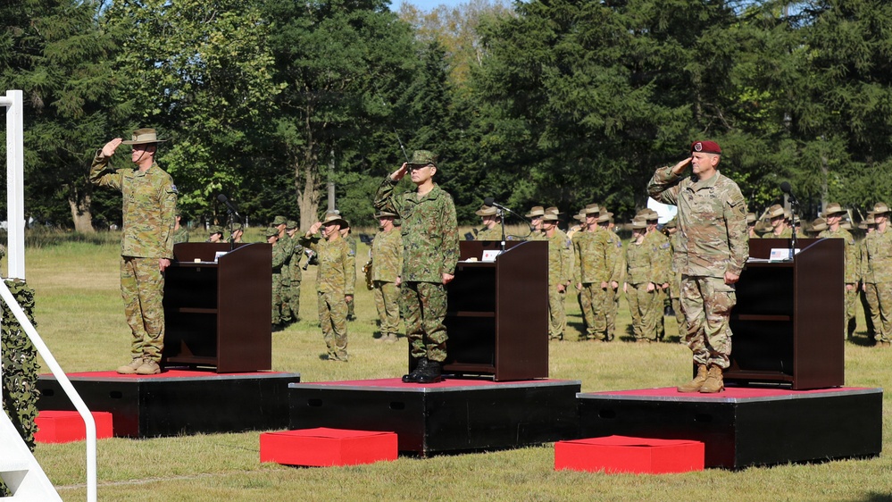 A formal military ceremony is taking place outdoors on a grassy field, featuring three senior officers standing on elevated platforms and rendering salutes simultaneously. The officers appear to represent at least two different nations based on their distinct uniforms and headgear. A large formation of troops stands at attention in the background.