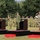 US and Japan Showcase Strong Partnership at Orient Shield 2026 Opening Ceremony A formal military ceremony is taking place outdoors on a grassy field, featuring three senior officers standing on elevated platforms and rendering salutes simultaneously. The officers appear to represent at least two different nations based on their distinct uniforms and headgear. A large formation of troops stands at attention in the background.