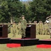 A formal military ceremony is taking place outdoors on a grassy field, featuring three senior officers standing on elevated platforms and rendering salutes simultaneously. The officers appear to represent at least two different nations based on their distinct uniforms and headgear. A large formation of troops stands at attention in the background.