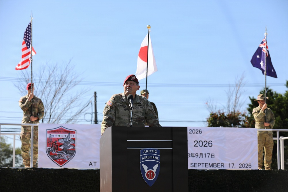 A U.S. Army officer wearing a red beret speaks at a podium bearing the 11th Airborne Division ('Arctic Airborne') insignia during an official military ceremony dated September 17, 2026. Three national flags — United States, Japan, and Australia — are displayed in the background, indicating a multinational military event. A banner partially visible behind the speaker references the same date in both English and Japanese.