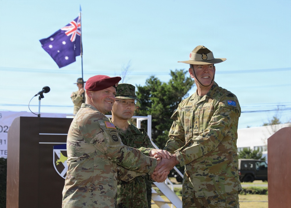 A military ceremony featuring three uniformed personnel from different nations shaking hands at what appears to be a formal event or exercise. An Australian flag is visible in the background, and the soldiers wear distinct uniforms representing the United States Army, a Japanese or Asian military force, and the Australian Army. The setting appears to be an outdoor military installation.