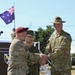 A military ceremony featuring three uniformed personnel from different nations shaking hands at what appears to be a formal event or exercise. An Australian flag is visible in the background, and the soldiers wear distinct uniforms representing the United States Army, a Japanese or Asian military force, and the Australian Army. The setting appears to be an outdoor military installation.