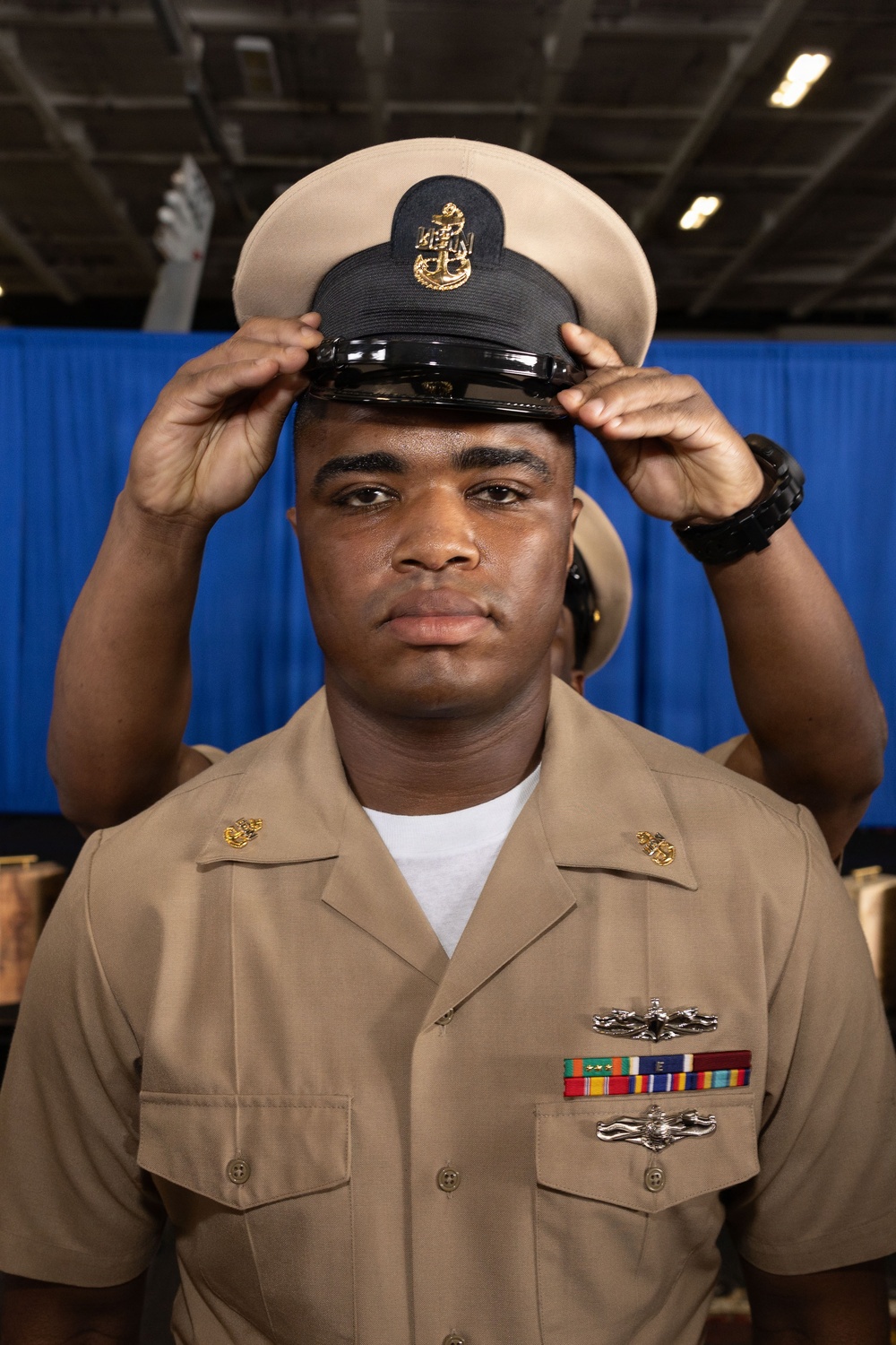 A U.S. Navy Chief Petty Officer is placing or adjusting his combination cover (cap) on his head during what appears to be a Chief Petty Officer pinning or frocking ceremony. He is wearing the khaki service uniform with visible warfare devices, service ribbons, and collar insignia. The blue draped backdrop and indoor setting suggest a formal military ceremony.