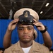 A U.S. Navy Chief Petty Officer is placing or adjusting his combination cover (cap) on his head during what appears to be a Chief Petty Officer pinning or frocking ceremony. He is wearing the khaki service uniform with visible warfare devices, service ribbons, and collar insignia. The blue draped backdrop and indoor setting suggest a formal military ceremony.