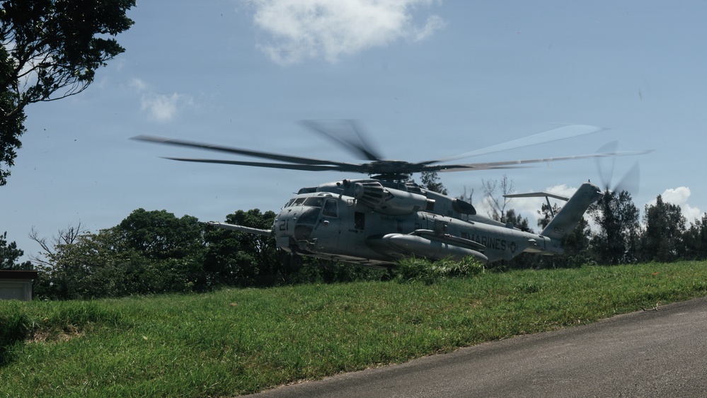 A U.S. Marines heavy-lift helicopter, identifiable as a Sikorsky CH-53E Super Stallion, is captured in motion with rotor blades visibly spinning and blurred. The aircraft appears to be either landing or taking off from a grassy area adjacent to a paved road, surrounded by dense tropical vegetation. The 'MARINES' marking is clearly visible on the fuselage.