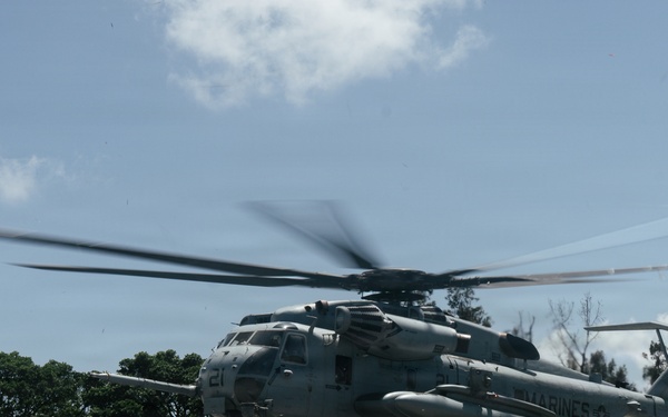 A U.S. Marines heavy-lift helicopter, identifiable as a Sikorsky CH-53E Super Stallion, is captured in motion with rotor blades visibly spinning and blurred. The aircraft appears to be either landing or taking off from a grassy area adjacent to a paved road, surrounded by dense tropical vegetation. The 'MARINES' marking is clearly visible on the fuselage.