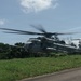 A U.S. Marines heavy-lift helicopter, identifiable as a Sikorsky CH-53E Super Stallion, is captured in motion with rotor blades visibly spinning and blurred. The aircraft appears to be either landing or taking off from a grassy area adjacent to a paved road, surrounded by dense tropical vegetation. The 'MARINES' marking is clearly visible on the fuselage.