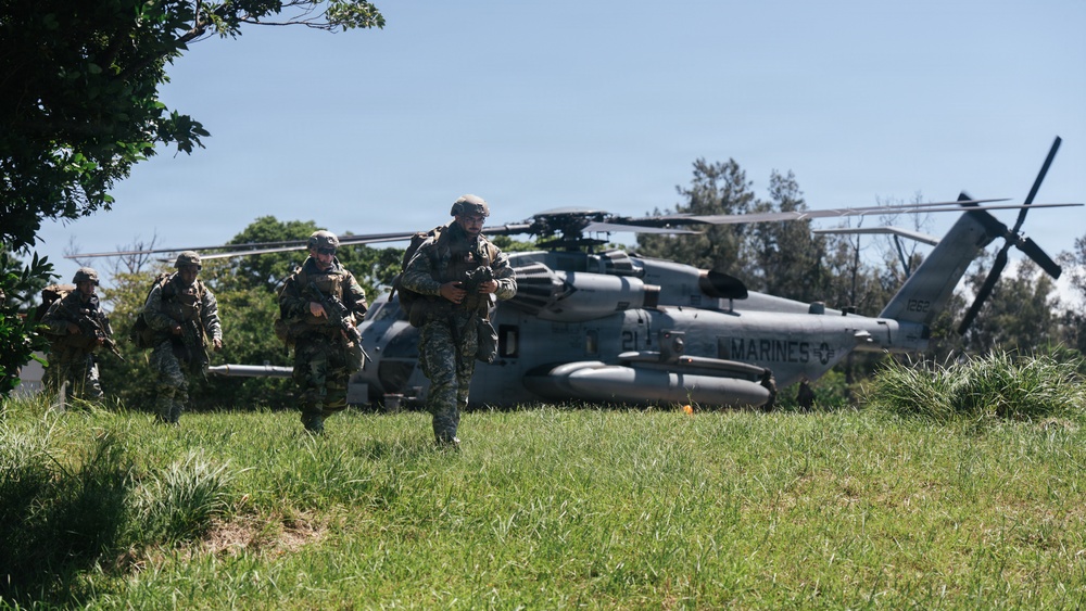 A group of armed military personnel in camouflage uniforms are moving across a grassy field away from a landed U.S. Marines helicopter. The helicopter, marked 'MARINES' with the number '21' visible on its fuselage, appears to be a CH-53E Super Stallion or similar heavy-lift helicopter. The scene appears to depict a tactical insertion or training exercise in a semi-rural environment.