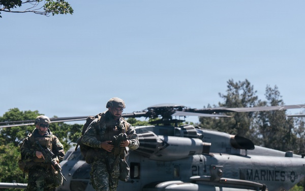 A group of armed military personnel in camouflage uniforms are moving across a grassy field away from a landed U.S. Marines helicopter. The helicopter, marked 'MARINES' with the number '21' visible on its fuselage, appears to be a CH-53E Super Stallion or similar heavy-lift helicopter. The scene appears to depict a tactical insertion or training exercise in a semi-rural environment.