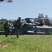 A group of armed military personnel in camouflage uniforms are moving across a grassy field away from a landed U.S. Marines helicopter. The helicopter, marked 'MARINES' with the number '21' visible on its fuselage, appears to be a CH-53E Super Stallion or similar heavy-lift helicopter. The scene appears to depict a tactical insertion or training exercise in a semi-rural environment.