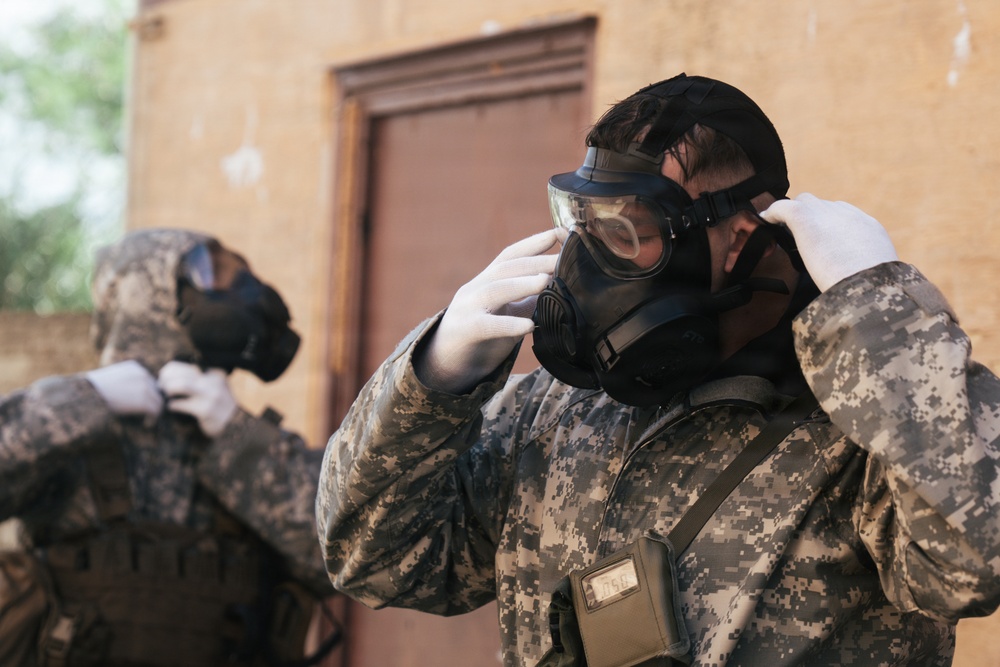 Two individuals wearing digital camouflage military uniforms are donning gas masks in front of a weathered building with a brown door. The foreground subject is actively adjusting their protective mask while wearing white gloves, with a device attached to their chest strap. The scene appears to depict a CBRN (Chemical, Biological, Radiological, Nuclear) drill or training exercise.