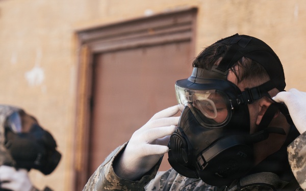 Two individuals wearing digital camouflage military uniforms are donning gas masks in front of a weathered building with a brown door. The foreground subject is actively adjusting their protective mask while wearing white gloves, with a device attached to their chest strap. The scene appears to depict a CBRN (Chemical, Biological, Radiological, Nuclear) drill or training exercise.