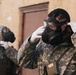 Two individuals wearing digital camouflage military uniforms are donning gas masks in front of a weathered building with a brown door. The foreground subject is actively adjusting their protective mask while wearing white gloves, with a device attached to their chest strap. The scene appears to depict a CBRN (Chemical, Biological, Radiological, Nuclear) drill or training exercise.