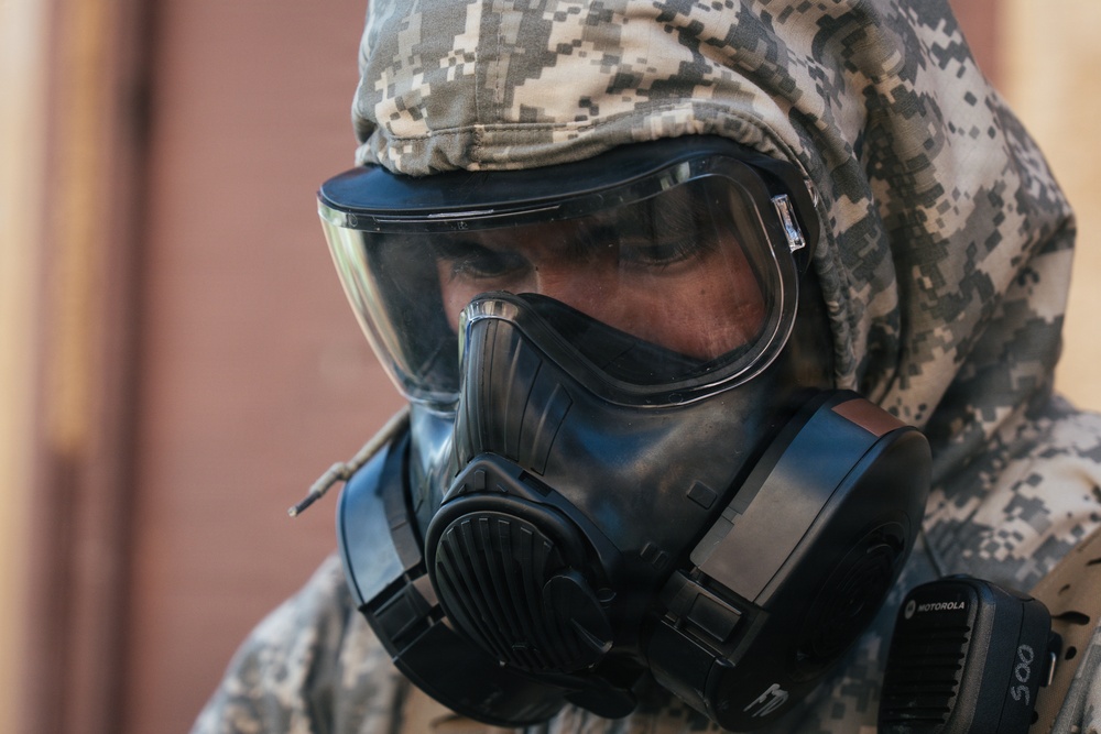 A person wearing military digital camouflage (ACU/UCP pattern) is equipped with a full-face gas mask and protective hood. The gear appears to be CBRN (Chemical, Biological, Radiological, Nuclear) protective equipment. A Motorola communication device is visible on the right side of the mask.