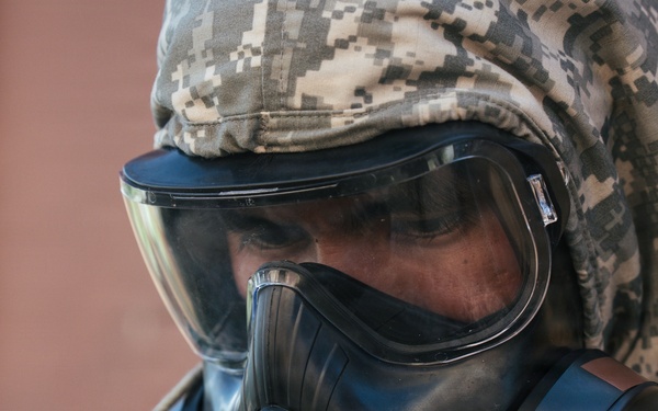 A person wearing military digital camouflage (ACU/UCP pattern) is equipped with a full-face gas mask and protective hood. The gear appears to be CBRN (Chemical, Biological, Radiological, Nuclear) protective equipment. A Motorola communication device is visible on the right side of the mask.