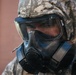 A person wearing military digital camouflage (ACU/UCP pattern) is equipped with a full-face gas mask and protective hood. The gear appears to be CBRN (Chemical, Biological, Radiological, Nuclear) protective equipment. A Motorola communication device is visible on the right side of the mask.