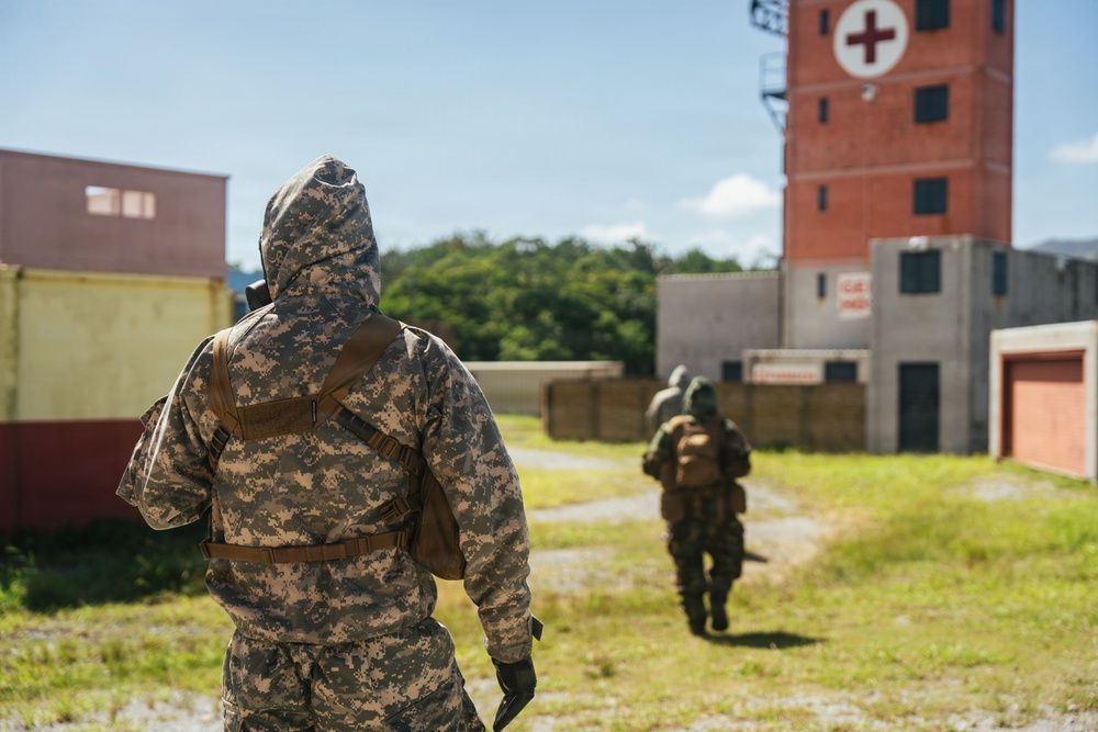 Two military personnel in full CBRN (Chemical, Biological, Radiological, Nuclear) protective gear and camouflage uniforms are moving through what appears to be a training facility or mock urban environment. The foreground soldier wears US Army Universal Camouflage Pattern (UCP) with a hooded protective suit and tactical backpack. A red brick tower marked with a Red Cross symbol is visible in the background, suggesting a medical or emergency response training scenario.
