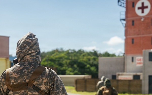 Two military personnel in full CBRN (Chemical, Biological, Radiological, Nuclear) protective gear and camouflage uniforms are moving through what appears to be a training facility or mock urban environment. The foreground soldier wears US Army Universal Camouflage Pattern (UCP) with a hooded protective suit and tactical backpack. A red brick tower marked with a Red Cross symbol is visible in the background, suggesting a medical or emergency response training scenario.