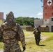 Two military personnel in full CBRN (Chemical, Biological, Radiological, Nuclear) protective gear and camouflage uniforms are moving through what appears to be a training facility or mock urban environment. The foreground soldier wears US Army Universal Camouflage Pattern (UCP) with a hooded protective suit and tactical backpack. A red brick tower marked with a Red Cross symbol is visible in the background, suggesting a medical or emergency response training scenario.