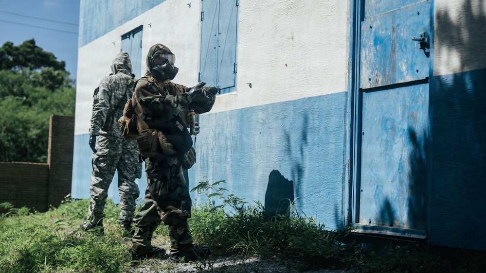 Two military personnel in full protective gear, including gas masks and camouflage uniforms, are conducting what appears to be a tactical training exercise near a blue and white painted building. The foreground figure is in a ready stance pointing toward a door, while the second figure stands nearby in standard ACU (Army Combat Uniform) pattern. The setting appears to be a training facility with overgrown vegetation along the building's base.