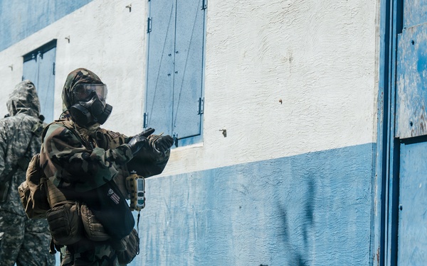 Two military personnel in full protective gear, including gas masks and camouflage uniforms, are conducting what appears to be a tactical training exercise near a blue and white painted building. The foreground figure is in a ready stance pointing toward a door, while the second figure stands nearby in standard ACU (Army Combat Uniform) pattern. The setting appears to be a training facility with overgrown vegetation along the building's base.