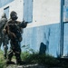 Two military personnel in full protective gear, including gas masks and camouflage uniforms, are conducting what appears to be a tactical training exercise near a blue and white painted building. The foreground figure is in a ready stance pointing toward a door, while the second figure stands nearby in standard ACU (Army Combat Uniform) pattern. The setting appears to be a training facility with overgrown vegetation along the building's base.