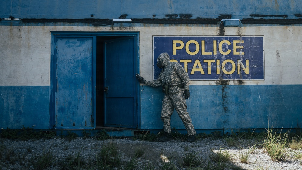 A person wearing military camouflage clothing and a gas mask is entering or inspecting an abandoned, weathered police station building. The building exterior is painted blue and white with a large 'POLICE STATION' sign mounted on the wall. The scene suggests either a post-disaster response, military exercise, or hazmat inspection scenario.