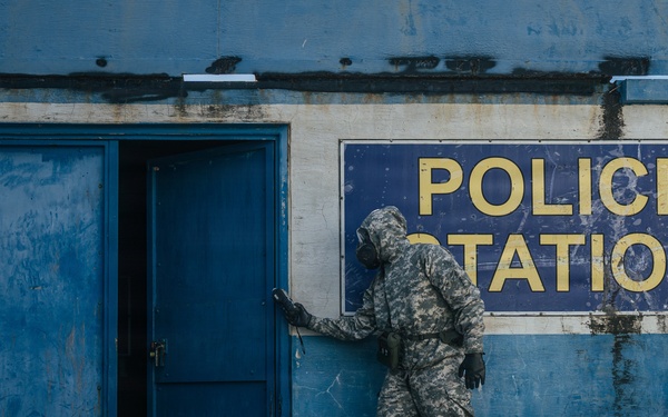 A person wearing military camouflage clothing and a gas mask is entering or inspecting an abandoned, weathered police station building. The building exterior is painted blue and white with a large 'POLICE STATION' sign mounted on the wall. The scene suggests either a post-disaster response, military exercise, or hazmat inspection scenario.