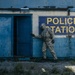 A person wearing military camouflage clothing and a gas mask is entering or inspecting an abandoned, weathered police station building. The building exterior is painted blue and white with a large 'POLICE STATION' sign mounted on the wall. The scene suggests either a post-disaster response, military exercise, or hazmat inspection scenario.