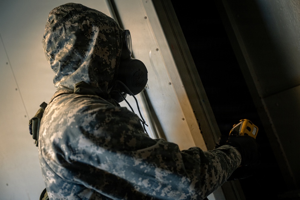 A person wearing US Army digital camouflage (ACU pattern) and a military-grade gas mask is holding a yellow handheld detection device near a metal surface. The individual appears to be conducting a CBRN (Chemical, Biological, Radiological, Nuclear) detection or inspection activity. The image is taken from a low angle, emphasizing the protective gear and the scanning action.
