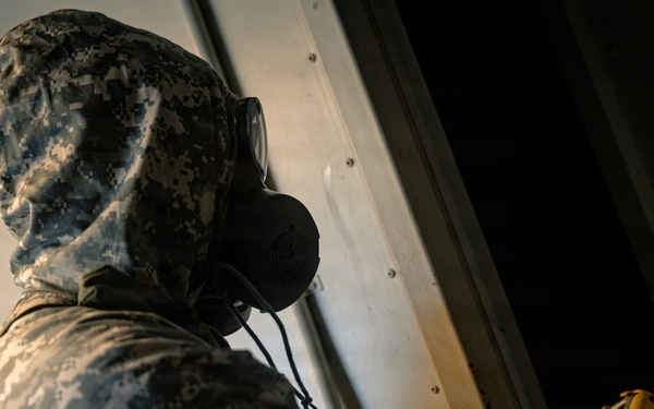 A person wearing US Army digital camouflage (ACU pattern) and a military-grade gas mask is holding a yellow handheld detection device near a metal surface. The individual appears to be conducting a CBRN (Chemical, Biological, Radiological, Nuclear) detection or inspection activity. The image is taken from a low angle, emphasizing the protective gear and the scanning action.
