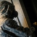 A person wearing US Army digital camouflage (ACU pattern) and a military-grade gas mask is holding a yellow handheld detection device near a metal surface. The individual appears to be conducting a CBRN (Chemical, Biological, Radiological, Nuclear) detection or inspection activity. The image is taken from a low angle, emphasizing the protective gear and the scanning action.