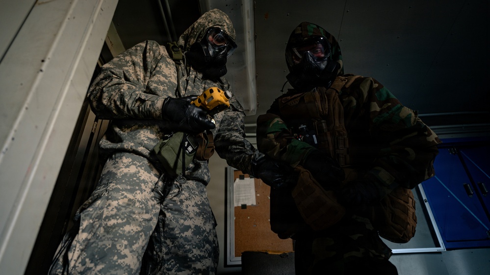 Two military personnel wearing full CBRN (Chemical, Biological, Radiological, and Nuclear) protective gear, including gas masks and camouflage suits, are engaged in what appears to be a training exercise or drill. The individual on the left wears US Army ACU (Army Combat Uniform) digital camouflage, while the one on the right wears woodland pattern camouflage. They appear to be inside a building, with one person holding a yellow handheld device, possibly a detection instrument.