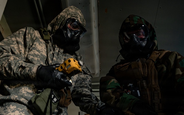 Two military personnel wearing full CBRN (Chemical, Biological, Radiological, and Nuclear) protective gear, including gas masks and camouflage suits, are engaged in what appears to be a training exercise or drill. The individual on the left wears US Army ACU (Army Combat Uniform) digital camouflage, while the one on the right wears woodland pattern camouflage. They appear to be inside a building, with one person holding a yellow handheld device, possibly a detection instrument.