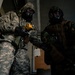 Two military personnel wearing full CBRN (Chemical, Biological, Radiological, and Nuclear) protective gear, including gas masks and camouflage suits, are engaged in what appears to be a training exercise or drill. The individual on the left wears US Army ACU (Army Combat Uniform) digital camouflage, while the one on the right wears woodland pattern camouflage. They appear to be inside a building, with one person holding a yellow handheld device, possibly a detection instrument.