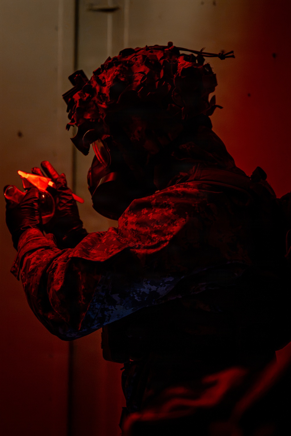 A heavily equipped military or tactical operator is photographed in near-darkness under intense red ambient lighting. The individual is wearing a camouflage helmet with foliage/ghillie attachments and what appears to be night-vision device mounts, along with tactical body armor. They are holding a glowing red chemlight (chemical light stick) in their gloved hand.