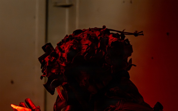 A heavily equipped military or tactical operator is photographed in near-darkness under intense red ambient lighting. The individual is wearing a camouflage helmet with foliage/ghillie attachments and what appears to be night-vision device mounts, along with tactical body armor. They are holding a glowing red chemlight (chemical light stick) in their gloved hand.