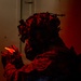 A heavily equipped military or tactical operator is photographed in near-darkness under intense red ambient lighting. The individual is wearing a camouflage helmet with foliage/ghillie attachments and what appears to be night-vision device mounts, along with tactical body armor. They are holding a glowing red chemlight (chemical light stick) in their gloved hand.