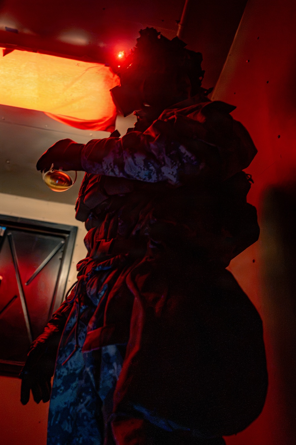 A figure wearing an elaborate, layered costume with ruffled fabric elements is photographed under dramatic red lighting. The person holds what appears to be a glass or goblet in one hand and wears long dark gloves. The low-key lighting creates a theatrical, moody atmosphere with the figure largely silhouetted against red-lit walls.
