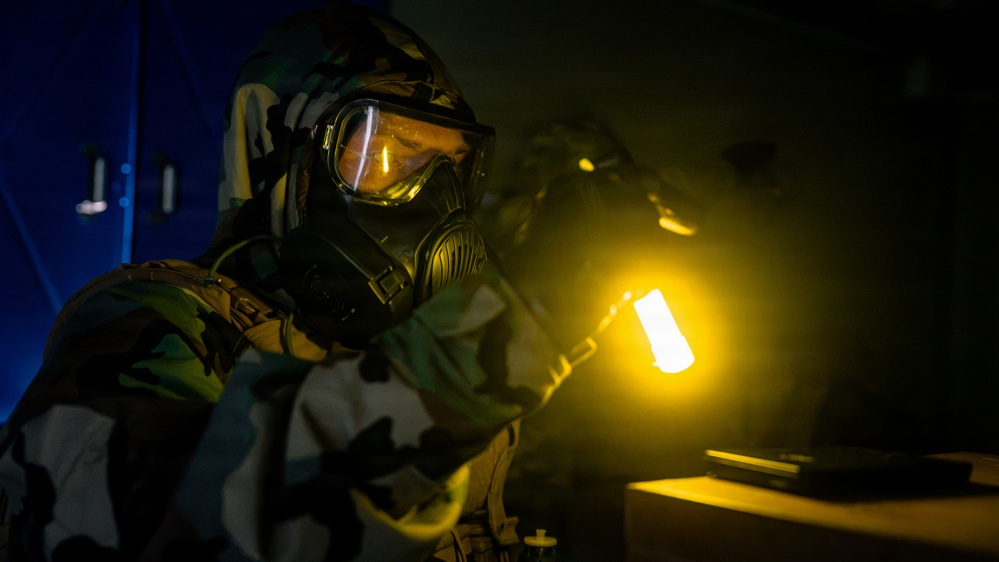 A person wearing full CBRN (Chemical, Biological, Radiological, Nuclear) protective gear, including a gas mask with wide-lens goggles and camouflage suit, is conducting an inspection or search in a darkened room. They are holding a bright yellow-glowing light source, likely a chemical light stick or tactical flashlight, which illuminates the immediate area. The scene suggests a military or emergency response training exercise or operational scenario.