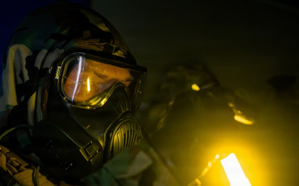 A person wearing full CBRN (Chemical, Biological, Radiological, Nuclear) protective gear, including a gas mask with wide-lens goggles and camouflage suit, is conducting an inspection or search in a darkened room. They are holding a bright yellow-glowing light source, likely a chemical light stick or tactical flashlight, which illuminates the immediate area. The scene suggests a military or emergency response training exercise or operational scenario.