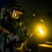A person wearing full CBRN (Chemical, Biological, Radiological, Nuclear) protective gear, including a gas mask with wide-lens goggles and camouflage suit, is conducting an inspection or search in a darkened room. They are holding a bright yellow-glowing light source, likely a chemical light stick or tactical flashlight, which illuminates the immediate area. The scene suggests a military or emergency response training exercise or operational scenario.