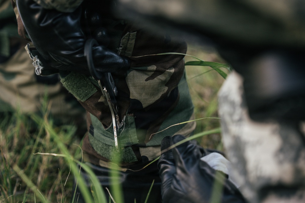 A close-up image showing a person wearing woodland camouflage clothing and black tactical gloves, appearing to handle a small metallic tool or instrument. The scene is set in an outdoor grassy environment. The focus is tight on the torso and hands area, with background elements blurred.