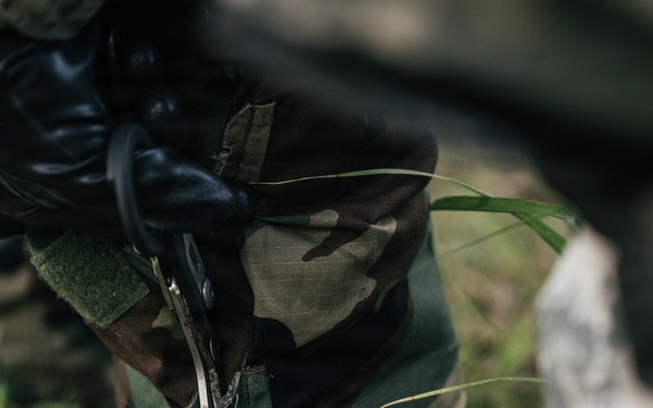 A close-up image showing a person wearing woodland camouflage clothing and black tactical gloves, appearing to handle a small metallic tool or instrument. The scene is set in an outdoor grassy environment. The focus is tight on the torso and hands area, with background elements blurred.