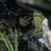 A close-up image showing a person wearing woodland camouflage clothing and black tactical gloves, appearing to handle a small metallic tool or instrument. The scene is set in an outdoor grassy environment. The focus is tight on the torso and hands area, with background elements blurred.