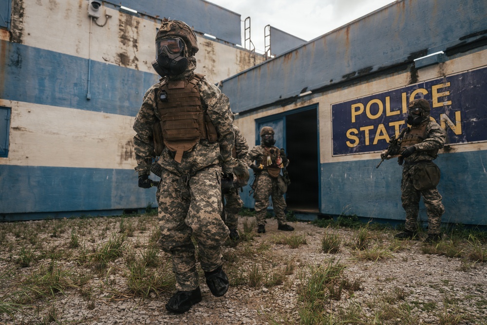 The image shows multiple armed personnel in camouflage uniforms and gas masks positioned outside a deteriorated building marked 'Police Station'. The structure shows significant weathering with blue and white paint, rust stains, and overgrown vegetation. The scene appears to depict a tactical military or law enforcement training exercise or simulation.