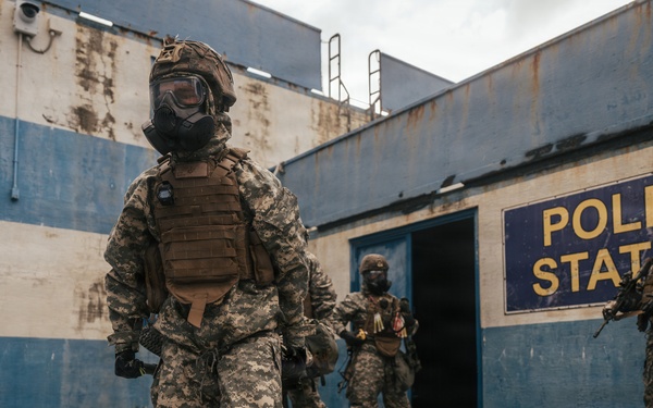 The image shows multiple armed personnel in camouflage uniforms and gas masks positioned outside a deteriorated building marked 'Police Station'. The structure shows significant weathering with blue and white paint, rust stains, and overgrown vegetation. The scene appears to depict a tactical military or law enforcement training exercise or simulation.