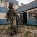 The image shows multiple armed personnel in camouflage uniforms and gas masks positioned outside a deteriorated building marked 'Police Station'. The structure shows significant weathering with blue and white paint, rust stains, and overgrown vegetation. The scene appears to depict a tactical military or law enforcement training exercise or simulation.