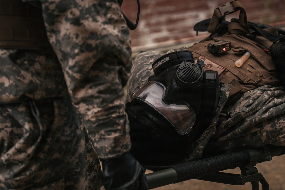 The image shows military equipment laid out on what appears to be a surface or rack, including a gas mask, tactical vest/plate carrier, and camouflage gear. A soldier in ACU (Army Combat Uniform) digital camouflage pattern is partially visible on the left side. The equipment appears to be staged or being prepared for use.