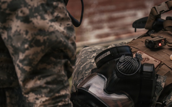 The image shows military equipment laid out on what appears to be a surface or rack, including a gas mask, tactical vest/plate carrier, and camouflage gear. A soldier in ACU (Army Combat Uniform) digital camouflage pattern is partially visible on the left side. The equipment appears to be staged or being prepared for use.
